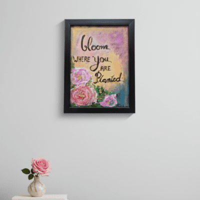 Framed inspirational quote artwork reading “Bloom where you are planted,” with painted flowers and soft pastel background, displayed on a minimalist wall.