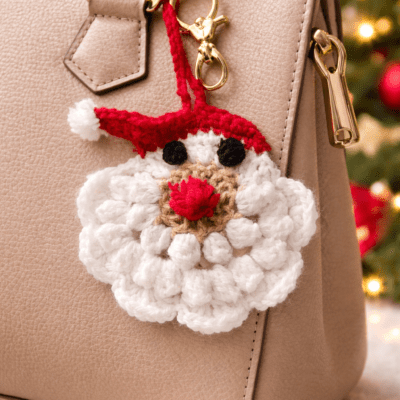 Handmade crochet Santa Claus bag charm with white beard and red hat hanging from a hand outdoors