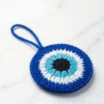 Handmade crochet evil eye charm in blue and white yarn with hanging loop on wooden background