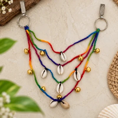 Handmade rainbow crochet cord hanging with cowrie shells and brass bells on dual keyrings against a marble background